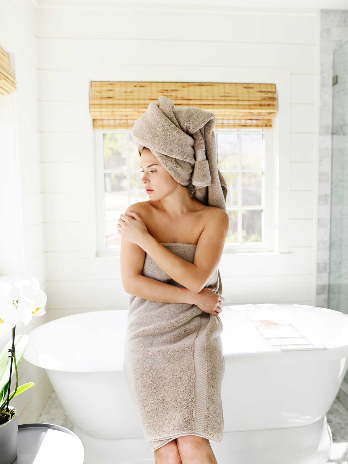 A woman sitting in the edge of a bathtub wrapped in a tan colored cotton bath towel.