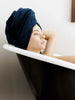 A woman laying in a bathtub with her hair up in a navy cotton bath towel.