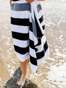 A woman walking barefoot along the shoreline of the beach, wrapped in a black, gray, and white striped cabana beach towel.