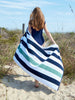 A woman walking on the beach while holding out a blue, green, and white striped cabana beach towel spread out behind her.