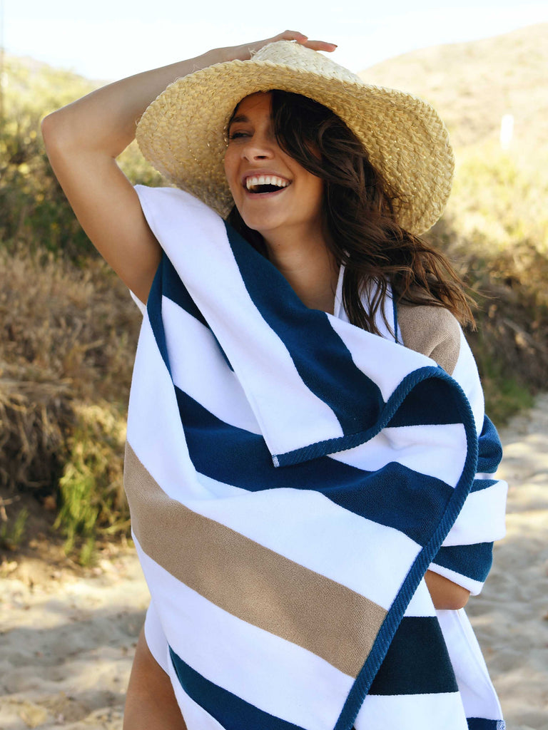 A woman standing on the beach, holding a hat on her head and wrapped in a blue, brown, and white striped cabana beach towel.