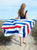 A woman walking on the beach while holding out a red, white, and blue striped cabana beach towel spread out behind her.