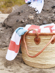 An orange, green, and white striped cabana beach towel in a beach bag sitting on the sand.