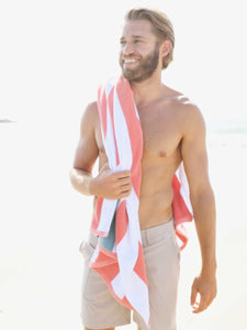 A man standing on the beach with an orange, green, and white striped cabana beach towel hanging over his shoulder. 