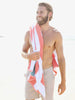 A man standing on the beach with an orange, green, and white striped cabana beach towel hanging over his shoulder. 