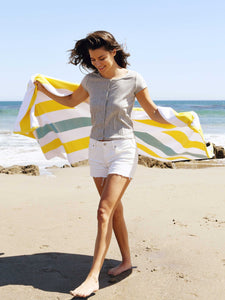 A woman walking on the beach while holding out a yellow, green, and white striped cabana beach towel spread out behind her.
