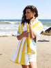 A woman standing on the beach with the ocean behind her, wrapped in a yellow, green, and white striped cabana beach towel.