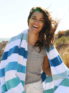 A woman on the beach wrapped in an extra large, striped cabana beach towel with shades of blue and green.