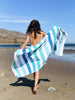 A woman walking on the beach while holding out a striped cabana beach towel with shades of blue and green behind her.