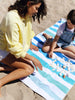 A woman and a girl sitting on a striped cabana beach towel with shades of blue and green while looking at seashells.