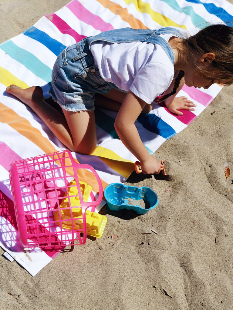 A girl sitting on a large, multicolor striped cabana beach towel while playing in the sand.