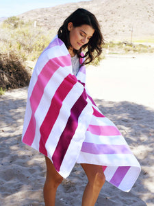 A woman standing in the sand with an extra large, pink, purple, and white striped cabana beach towel wrapped around her.