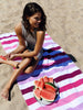 A woman sitting on a pink, purple, and white striped cabana beach towel in the sand while enjoying a piece of watermelon.