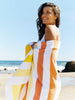 A woman in a white bathing suit on the beach wrapped in an extra large, yellow, orange, and white striped cabana beach towel.