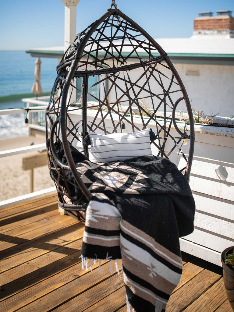 A black and Mexican blanket draped over a hanging char on a balcony overlooking the ocean.