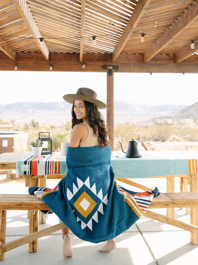 Woman in the desert sitting at table wrapped in navy and yellow Mexican blanket.