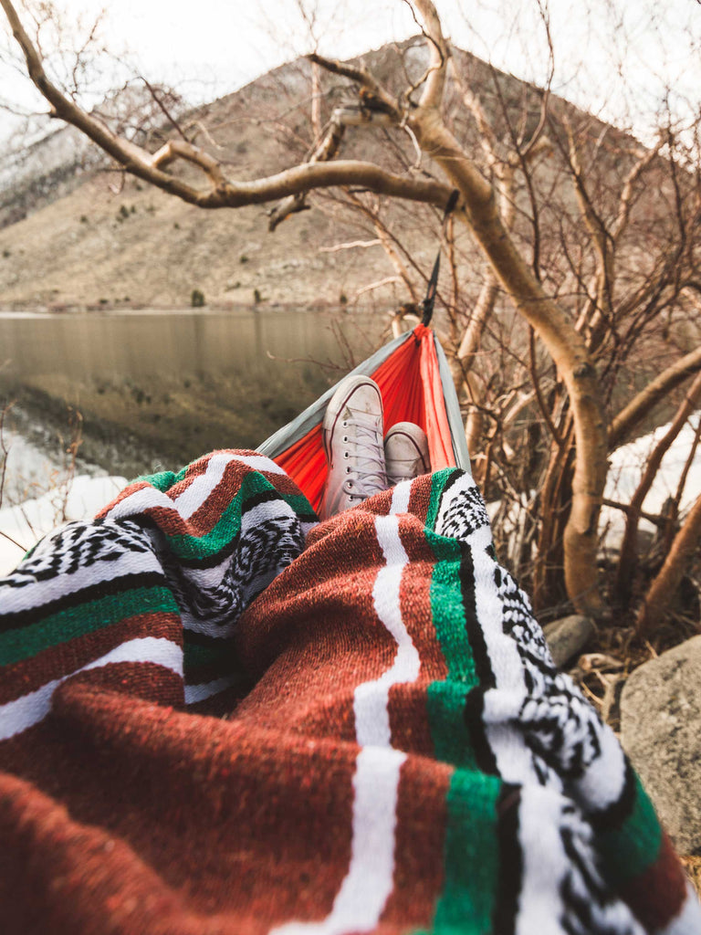 A person lying in a hammock on a lake using a green, red, and white Mexican blanket over their legs.