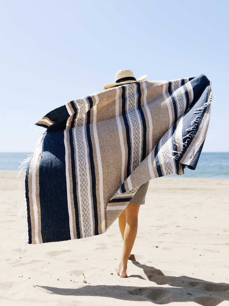 A woman walking on the beach holding a gray, tan, and white Mexican blanket spread out behind her.