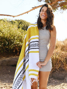 A woman holding a large, yellow and white striped Mexican blanket over her shoulder at the beach. 