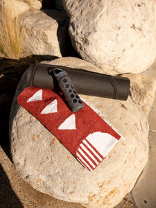 A folded Mexican blanket in a red pattern on a beach rock next to a black yoga mat and under a gray water bottle.