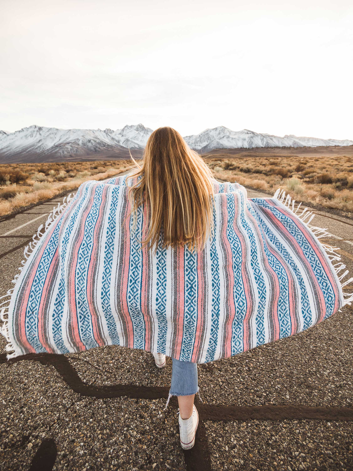 A woman walking on a road in a desert with a traditional blue, pink, and gray Mexican blanket wrapped around her. 