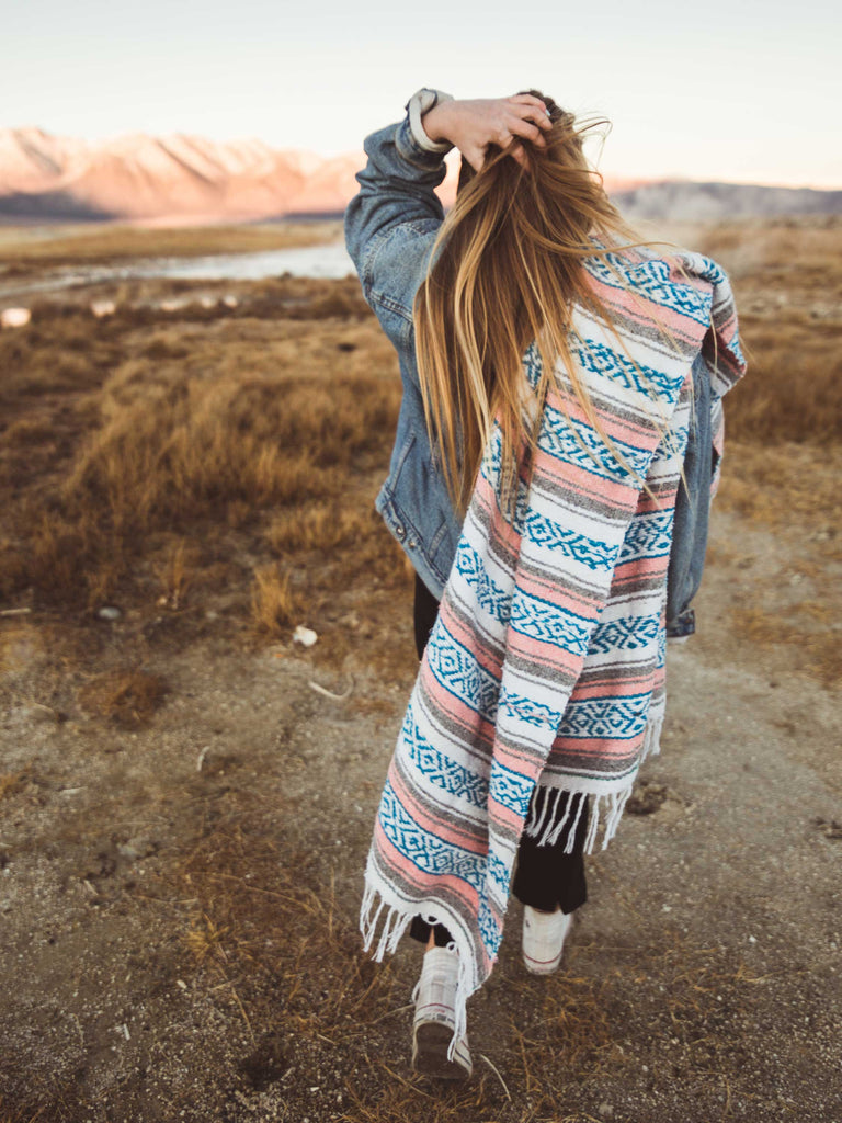 A woman walking in a desert with a traditional blue, pink, and gray Mexican blanket thrown over one of her shoulders.