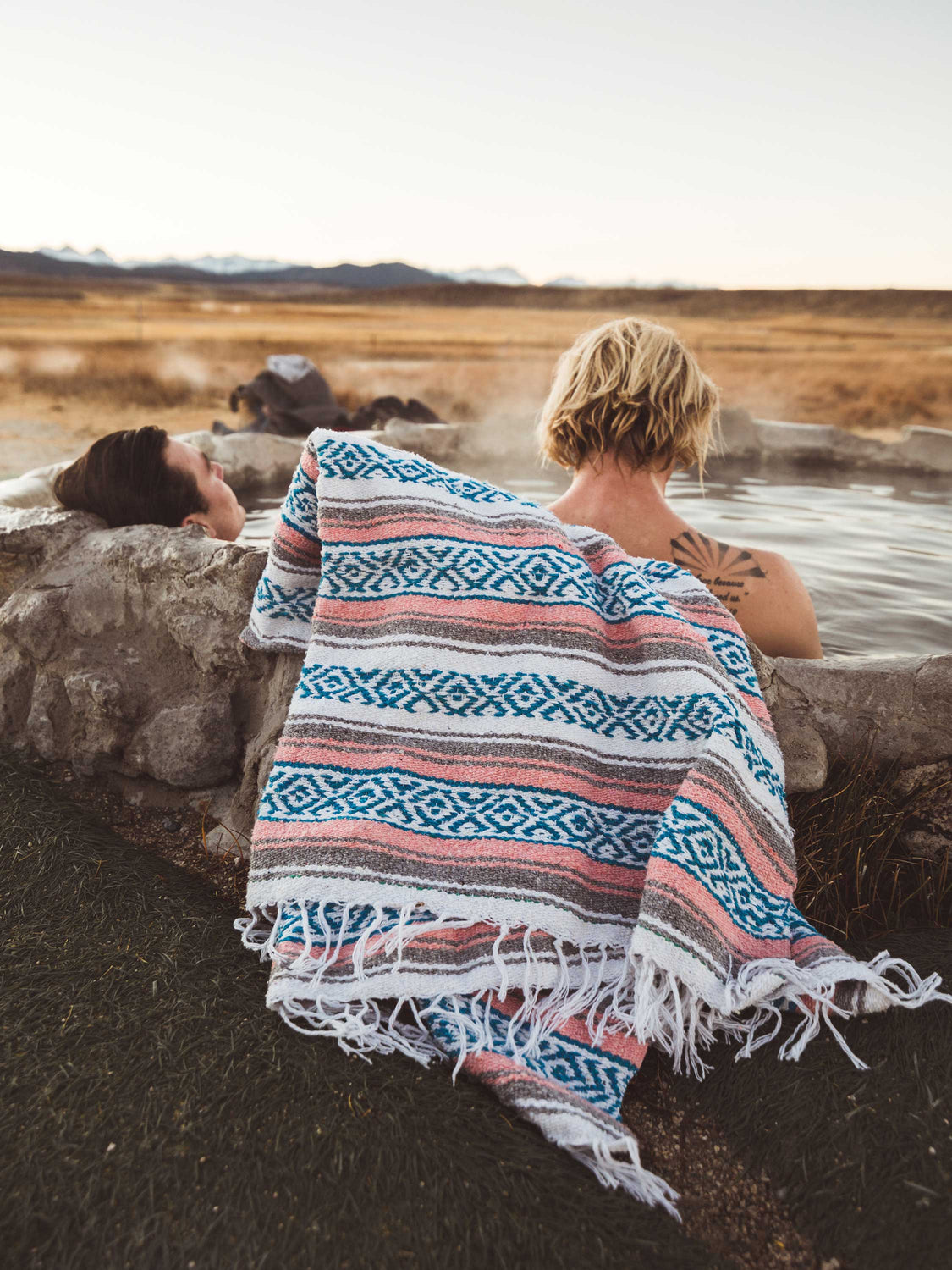 A man and a woman relaxing in a hot spring with a blue, pink, and gray traditional Mexican blanket draped over the side of the spring.