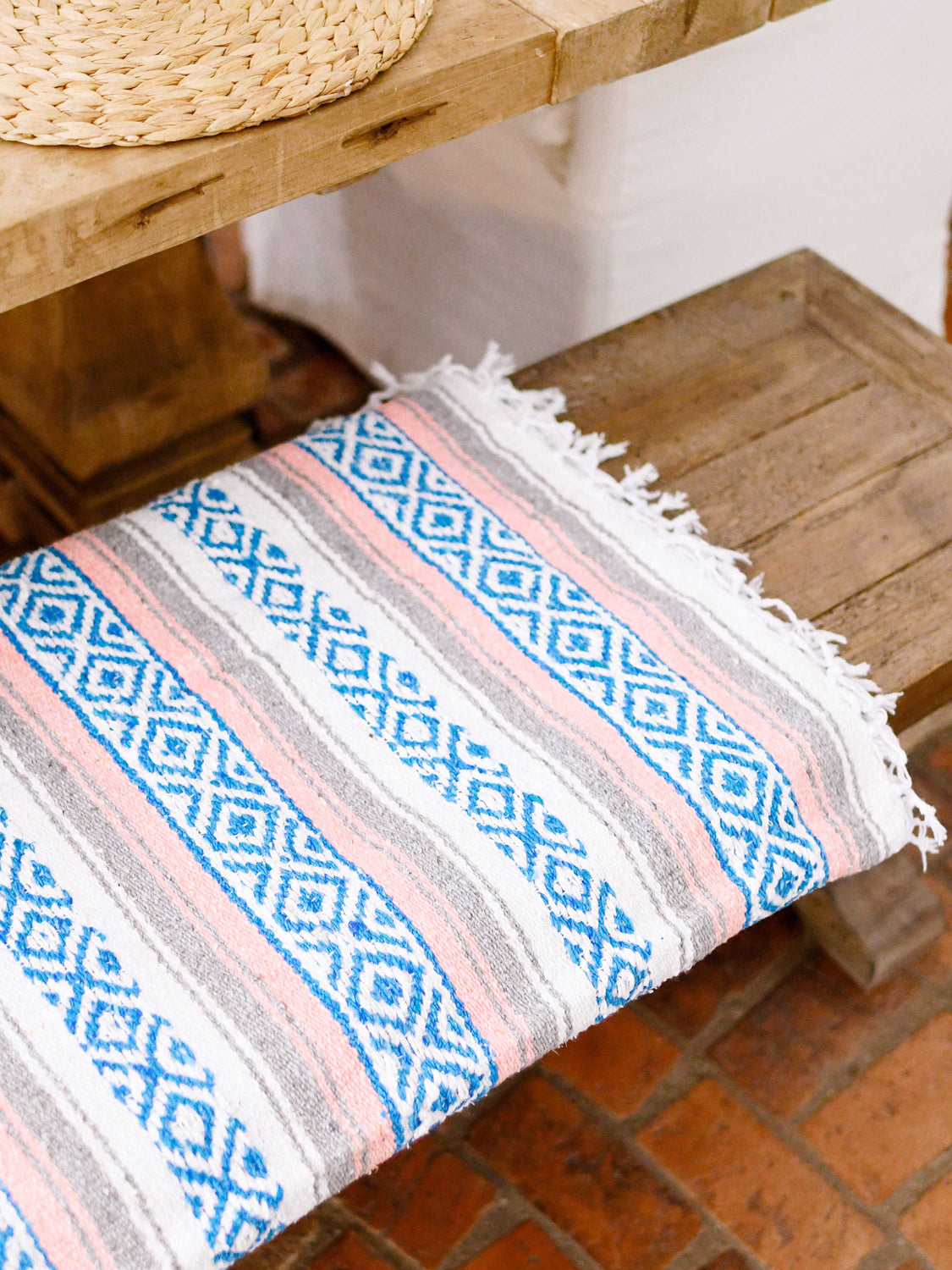 A blue, pink, and gray oversized Mexican blanket folded on a wooden bench. 