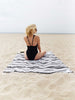 A woman sitting with her back turned on an oversized black, gray, and white Mexican blanket on the beach. 