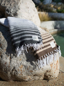A gray and white striped Mexican blanket folded on top of a brown, tan, and white striped Mexican blanket poolside.