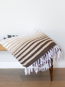 A hand-woven, brown striped Mexican blanket folded on top of a wicker bench. 