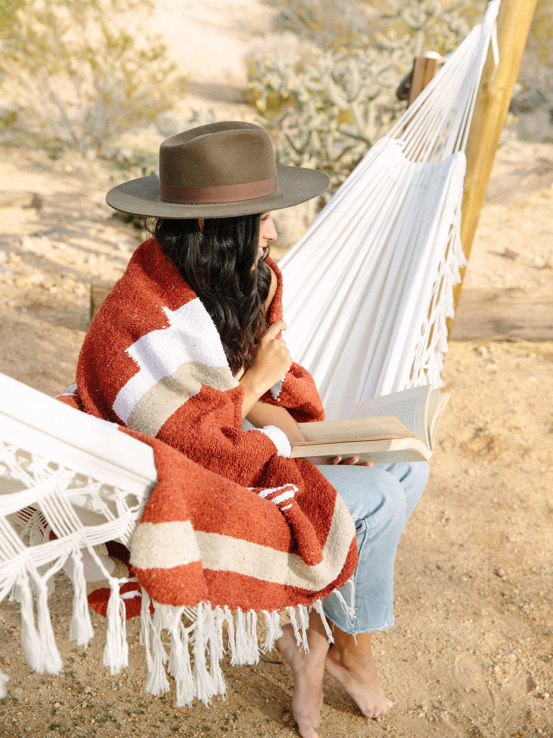 Woman sitting on hammock in desert wrapped in red and burnt orange Mexican blanket