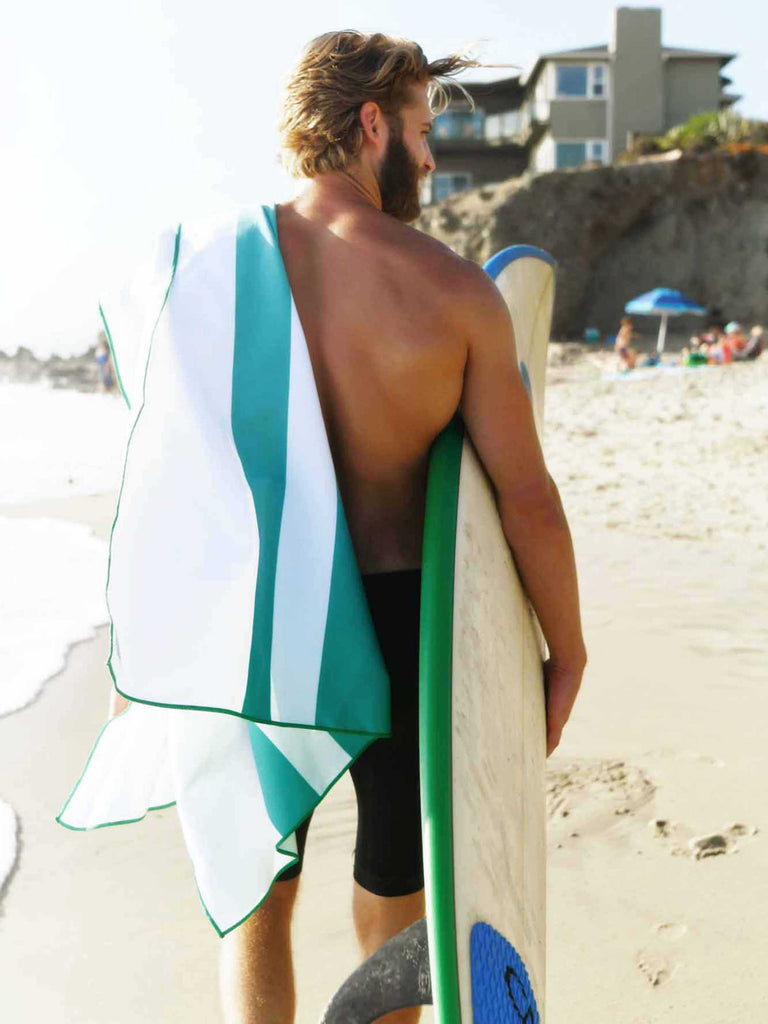 A man on the beach carrying a surfboard under his arm and an aqua blue striped microfiber beach towel over his shoulder.