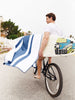 A man biking while holding his surfboard with a navy blue and white striped microfiber beach towel hanging over it.