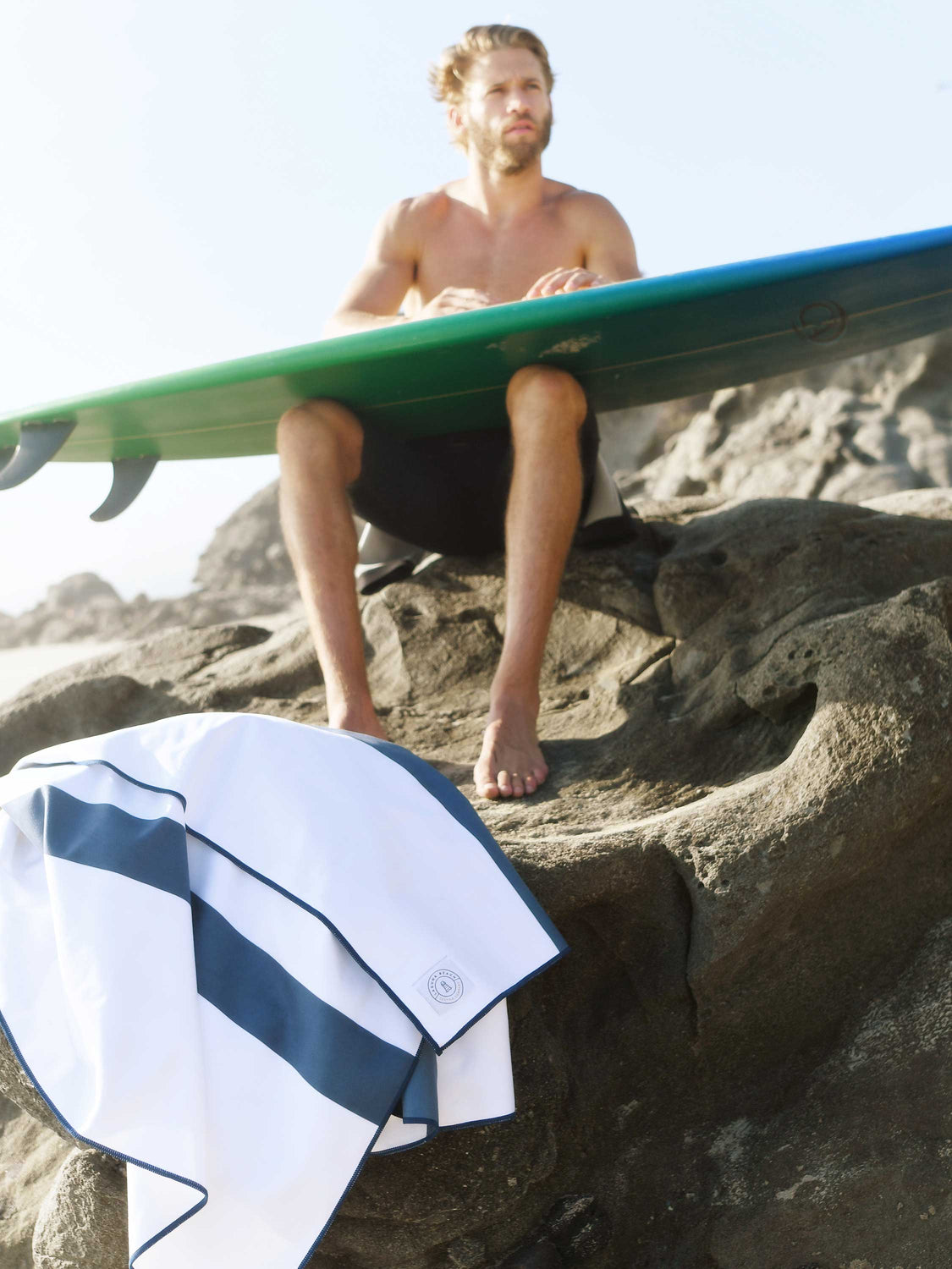 An oversized navy blue striped microfiber beach towel sitting at the foot of a rock near a man waxing his surfboard.
