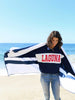 A woman wearing a sweatshirt that says ‘Laguna,’ holding a navy blue striped microfiber beach towel spread out behind her.