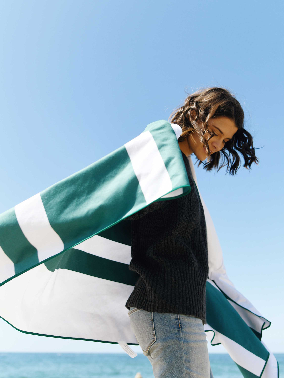 A woman wrapped in an oversized green and white striped microfiber beach towel blowing in the ocean breeze.