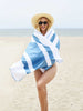 A woman in a bathing suit, hat, and sunglasses wrapped in a lightweight, oversized light blue striped microfiber beach towel.