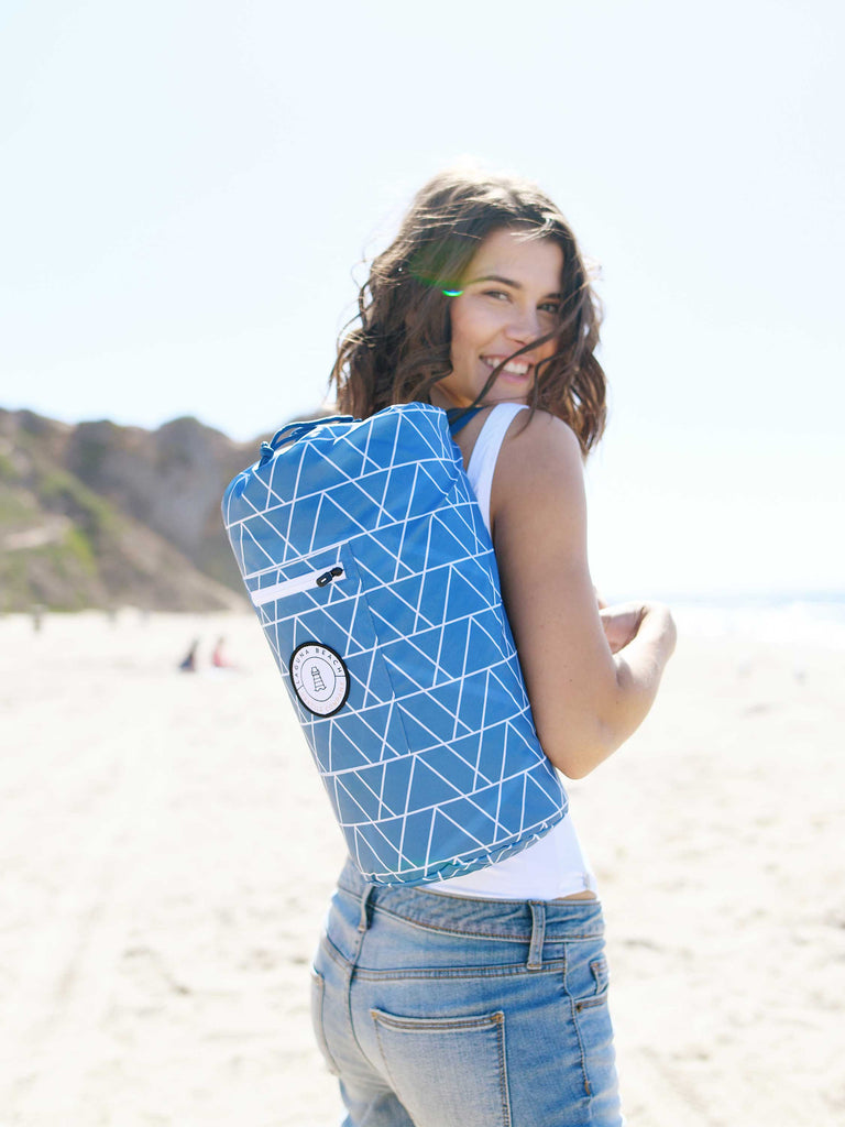 A woman on a beach carrying a small case packed with a waterproof, oversized blue picnic and outdoor blanket on her shoulder.