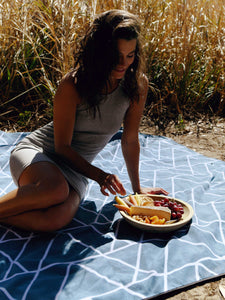 A woman enjoying a picnic on an oversized, blue and white mountain patterned waterproof outdoor blanket while camping.