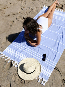 A woman in a white bathing suit laying out on an oversized blue and white striped Turkish towel at the beach.