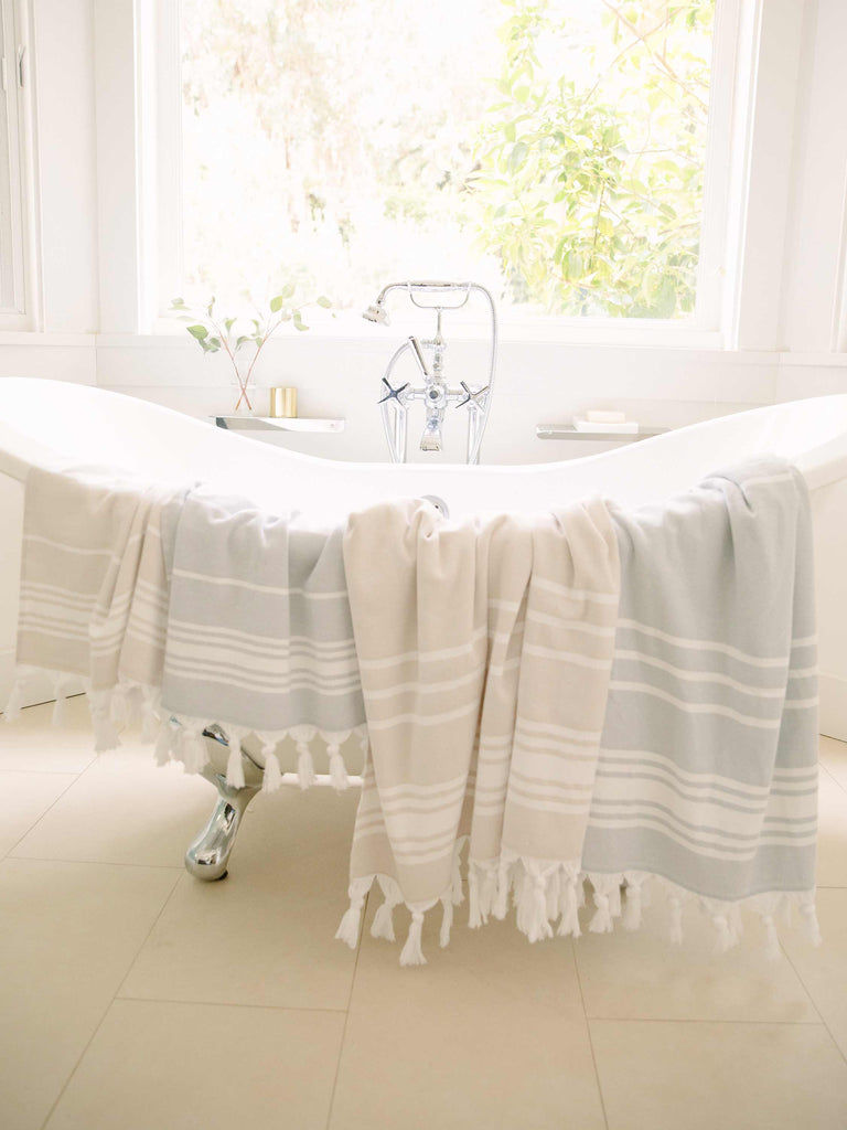 Four tan and white and charcoal and white striped hand and beach Turkish towels draped over a porcelain bathtub.