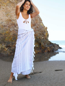 A woman standing in front of a large rock on the beach, wearing a gray and white striped Turkish beach towel as a sarong.