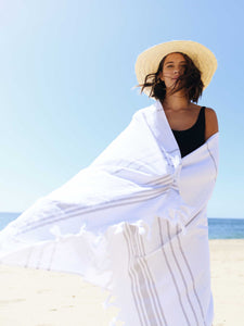 A woman standing on the beach wrapped in an extra large, white and gray striped Turkish towel blowing in the wind.