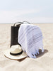 A white and gray striped Turkish towel in a black beach bag sitting on the sand next to a hat.