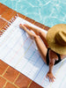 A woman sitting by the pool on an extra large, white and rainbow striped Turkish beach towel.