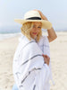 A woman standing on the beach while holding a hat on her head, wrapped in an oversized, white and gray striped Turkish towel.