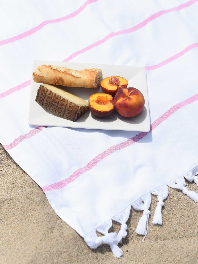 A charcuterie plate atop an oversized, white and pink striped Turkish beach towel laid out on the sand.