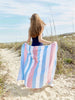 A woman standing on the beach with her back turned, holding a blue, pink, and white striped Turkish beach towel behind her.