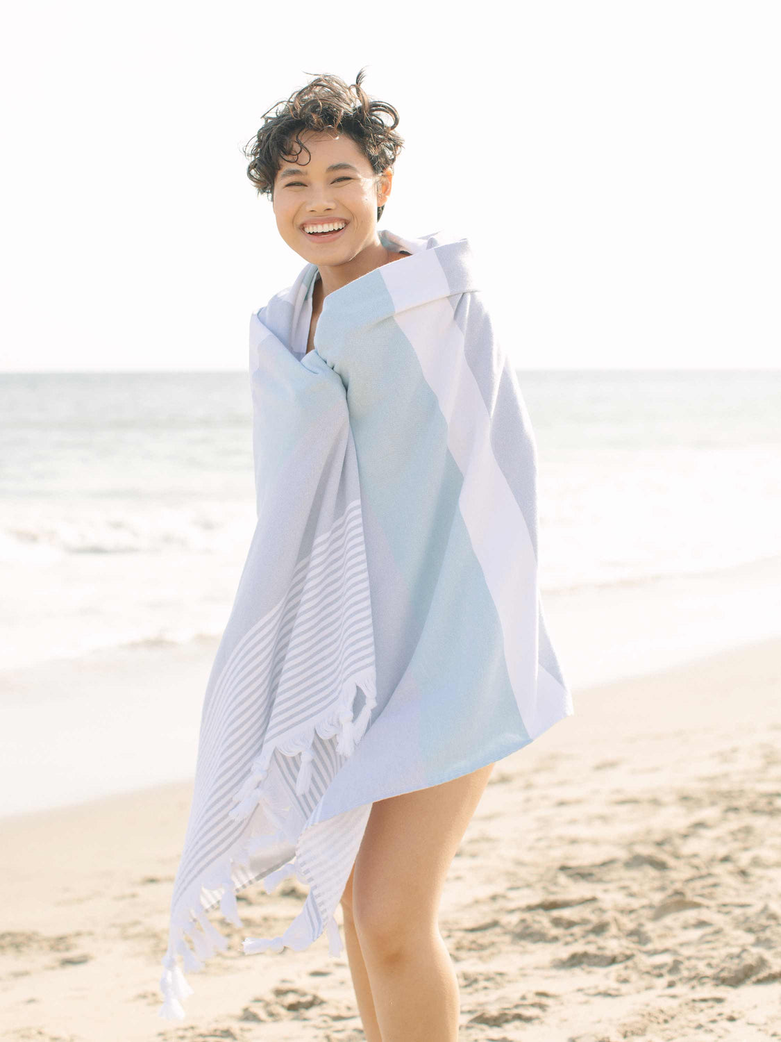 A woman wrapped in a teal and grey oversized Turkish towel smiling on the beach.